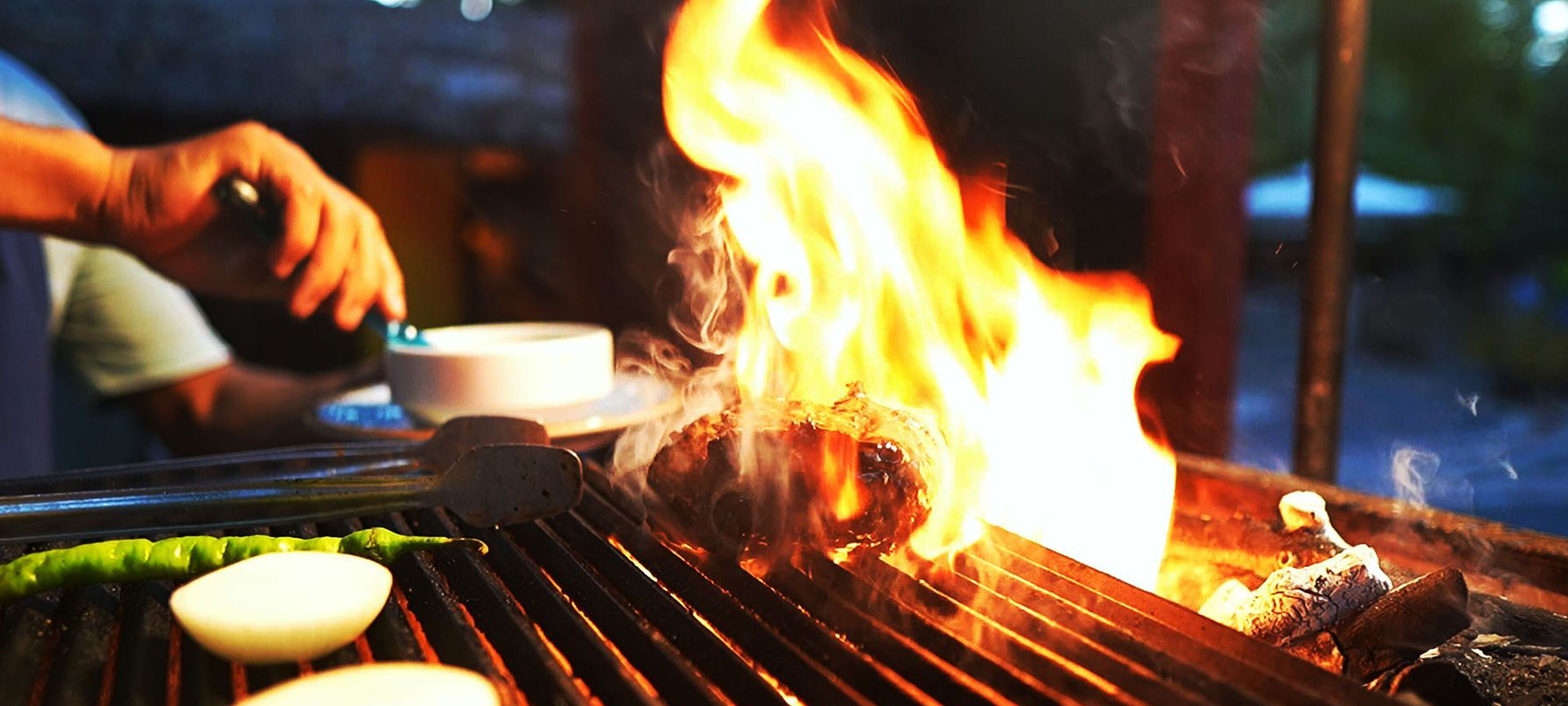 Una parrilla con comida cocinándose sobre fuego vivo, una persona asando y un recipiente con agua cerca.