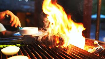 Una parrilla con comida cocinándose sobre fuego vivo, una persona asando y un recipiente con agua cerca.