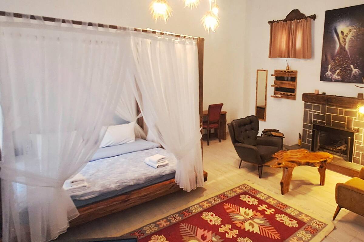 A bedroom with a four-poster bed, a fireplace, a chair, and a small table.