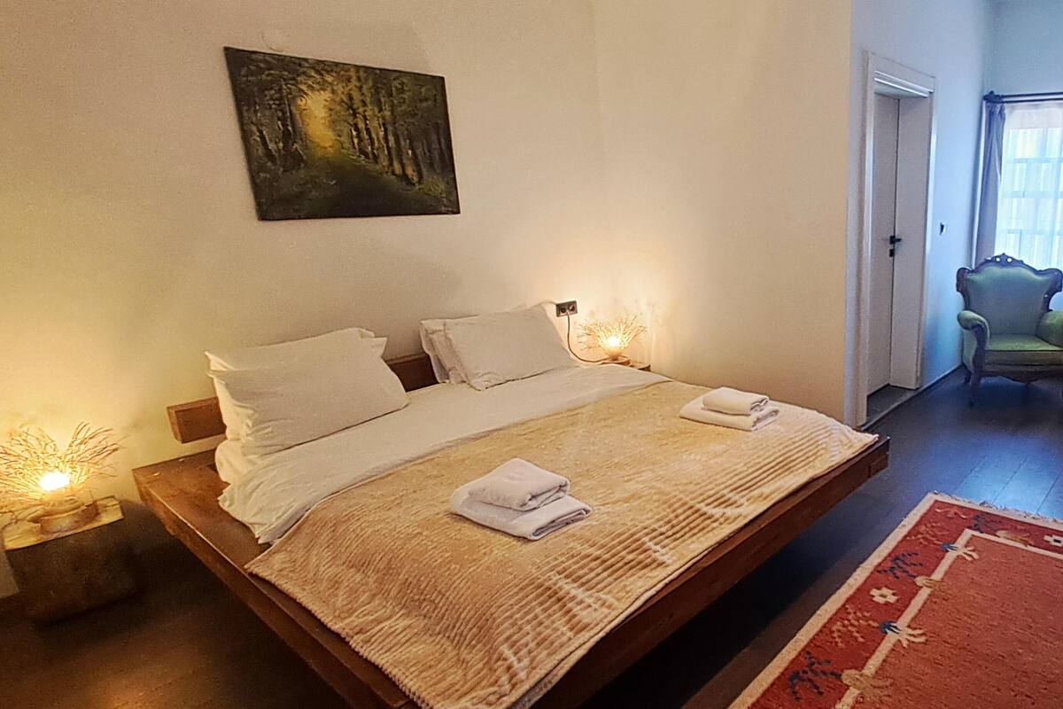 A bedroom with a bed, two bedside tables, a nightstand, a painting on the wall, and a chair in the corner.