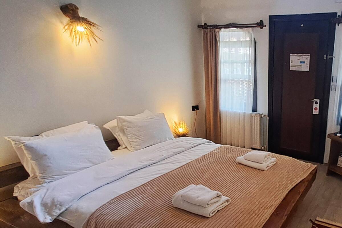 A neatly made bed with white linens and two folded towels, a wall-mounted light fixture, a window with curtains, and a door with a sign.