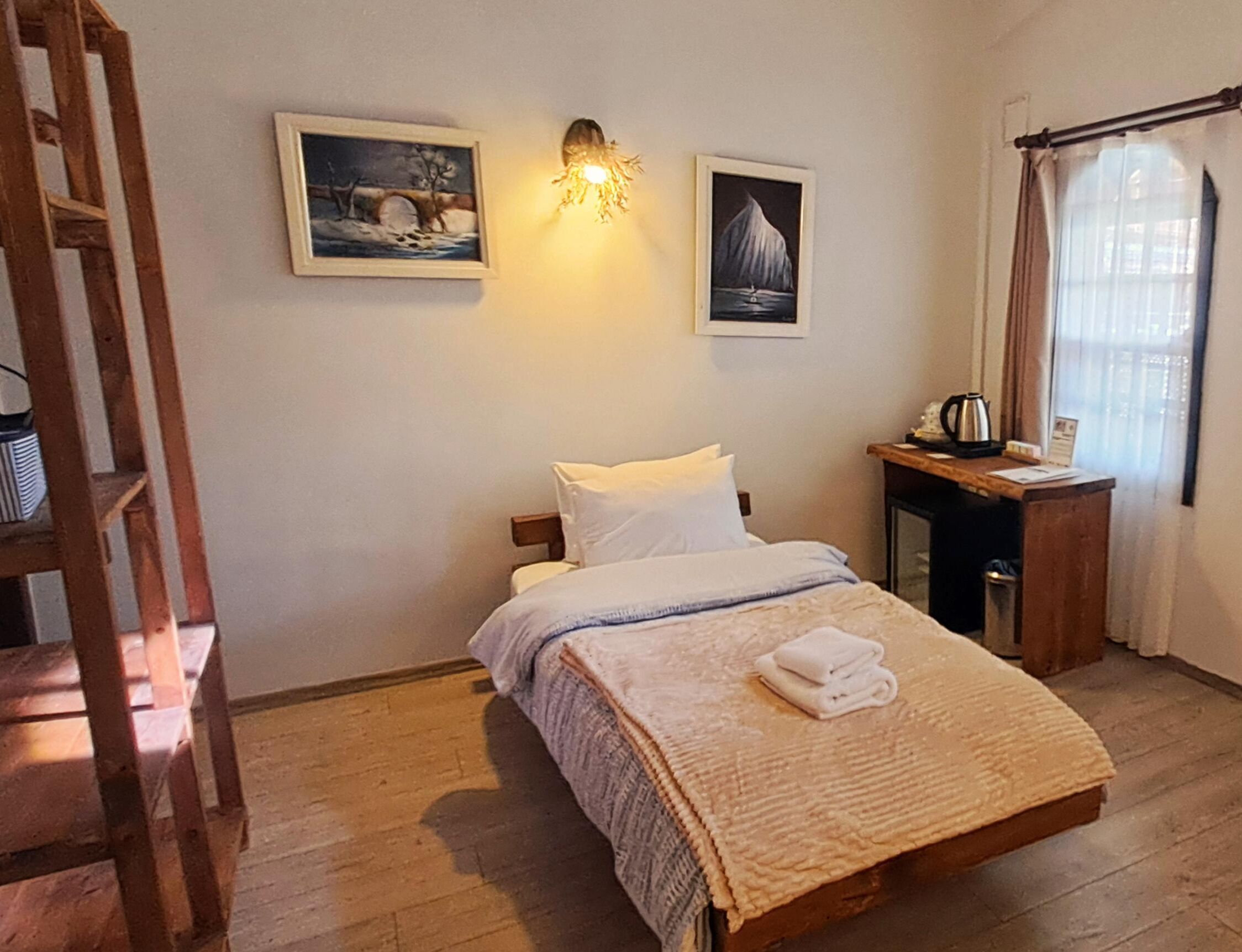 A bedroom with a bed, wooden floor, a desk with a kettle, and two framed pictures on the wall.