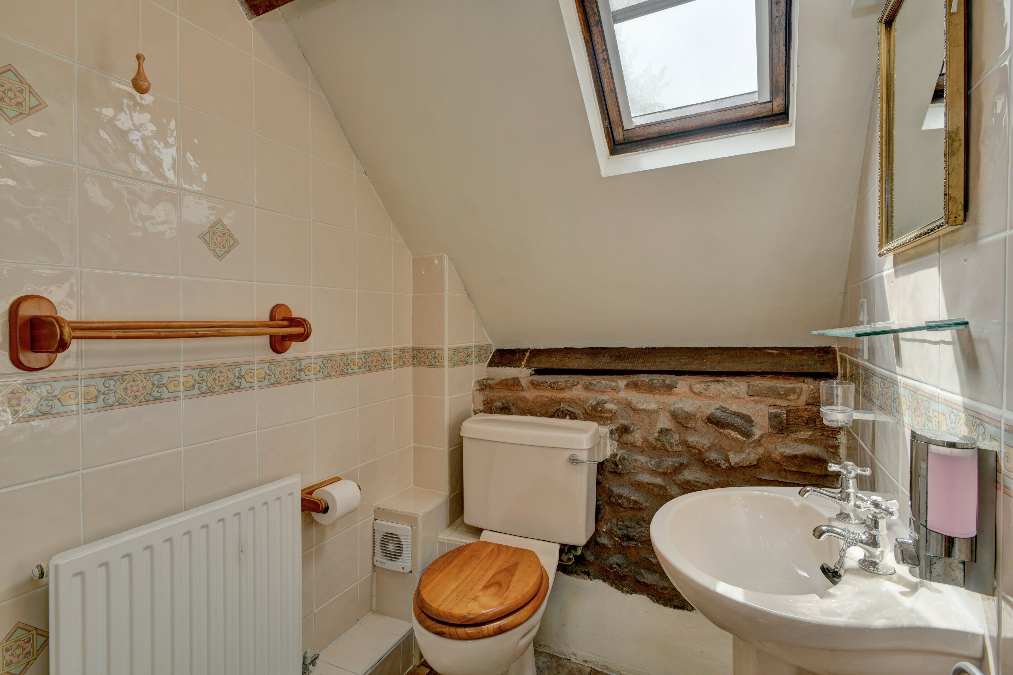 Bany amb vàter de fusta, un lavabo blanc, paret de pedra i una finestra.