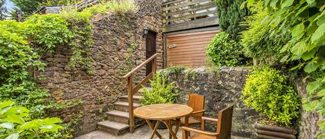 A stone building with a wooden staircase, a round wooden table with chairs, and a wooden fence.