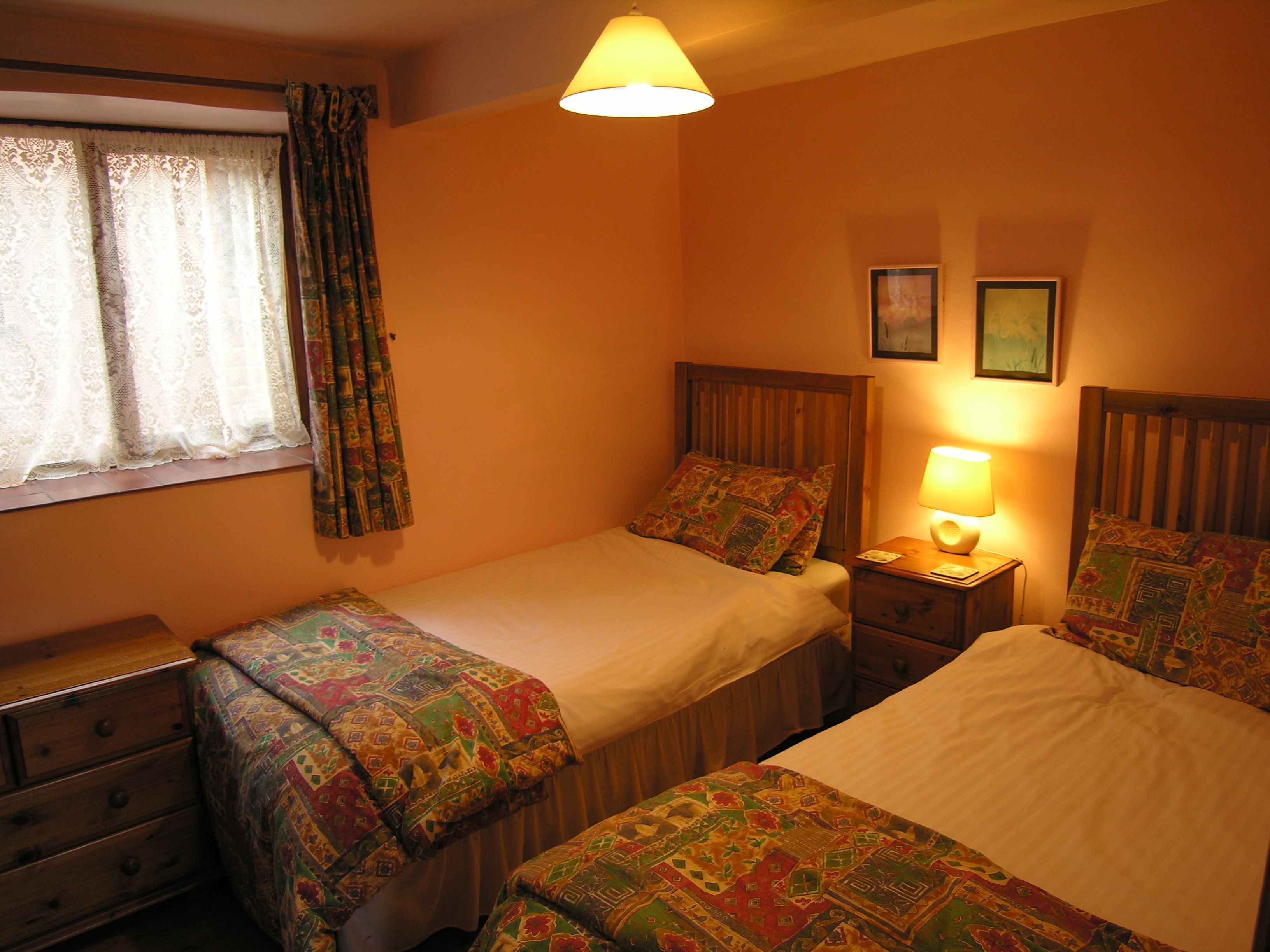 A bedroom with two beds, a window with curtains, a bedside lamp, and a wooden dresser.