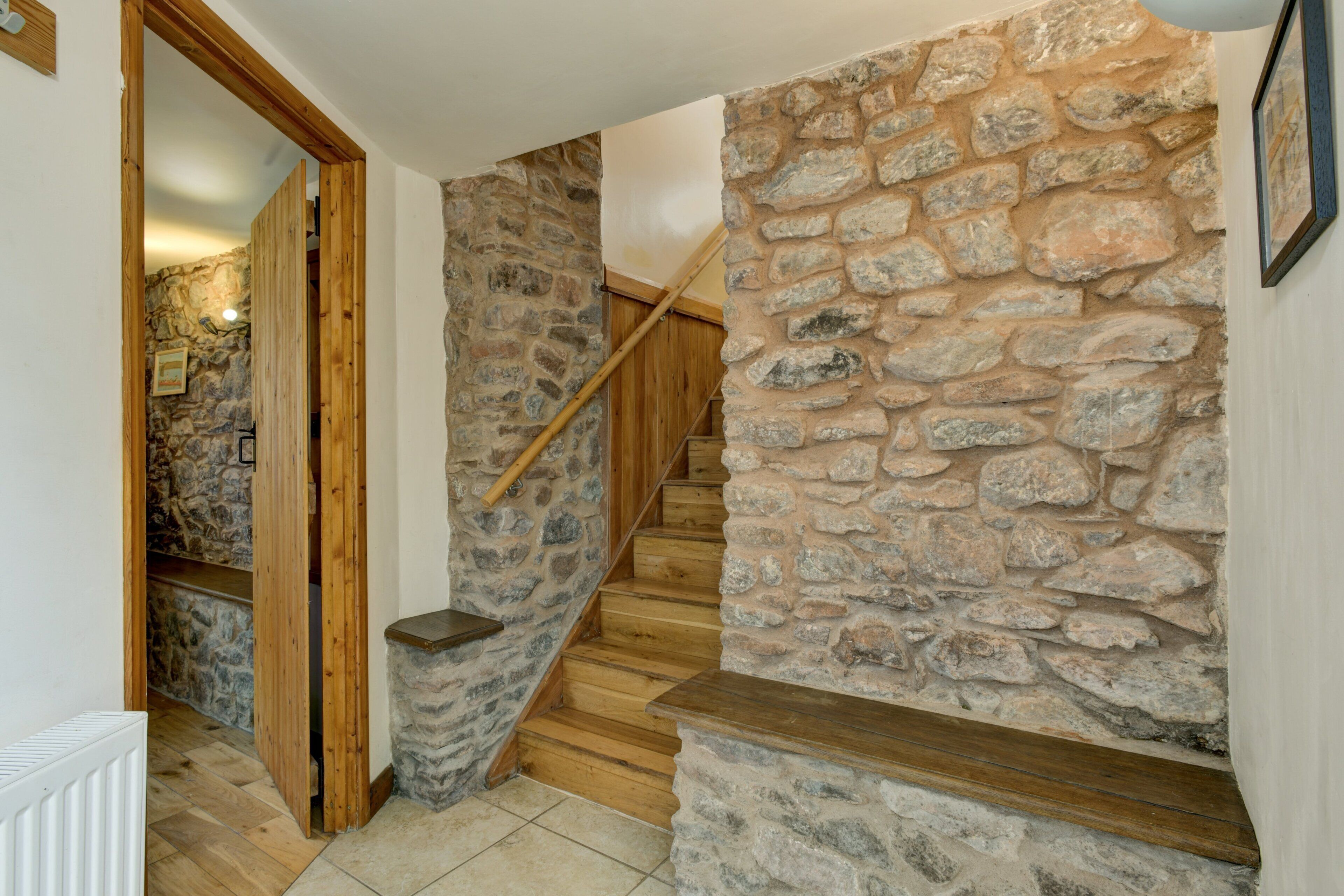 Una escalera de madera con paredes de piedra que conduce a una habitación con un radiador blanco y una puerta de madera.