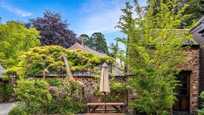 A rustic stone building with a thatched roof, surrounded by lush greenery and a gravel path. - Luccombe Cottage, Near Dunster (Near Dunster)