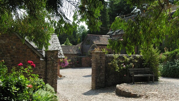 A stone building with a gravel path, a wooden bench, and a stone wall with flowers. - Luccombe Cottage, Near Dunster (Near Dunster)