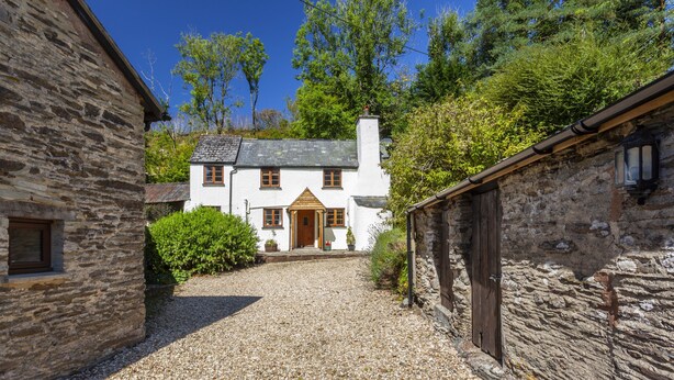 A stone building with a white facade, a wooden door, and a gravel driveway leading to a white house with a grey roof. - Hedgehog Cottage, Wheddon Cross (Wheddon Cross)
