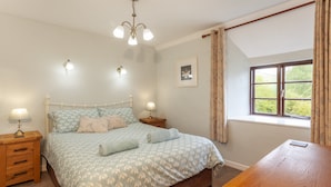A bedroom with a bed, bedside lamps, a wooden nightstand, a wooden table, a window with curtains, and a ceiling light fixture. - Hedgehog Cottage, Wheddon Cross (Wheddon Cross)