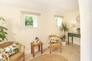 A room with two wicker chairs, a small wooden table with a tray, a potted plant, and a green desk with a lamp.
