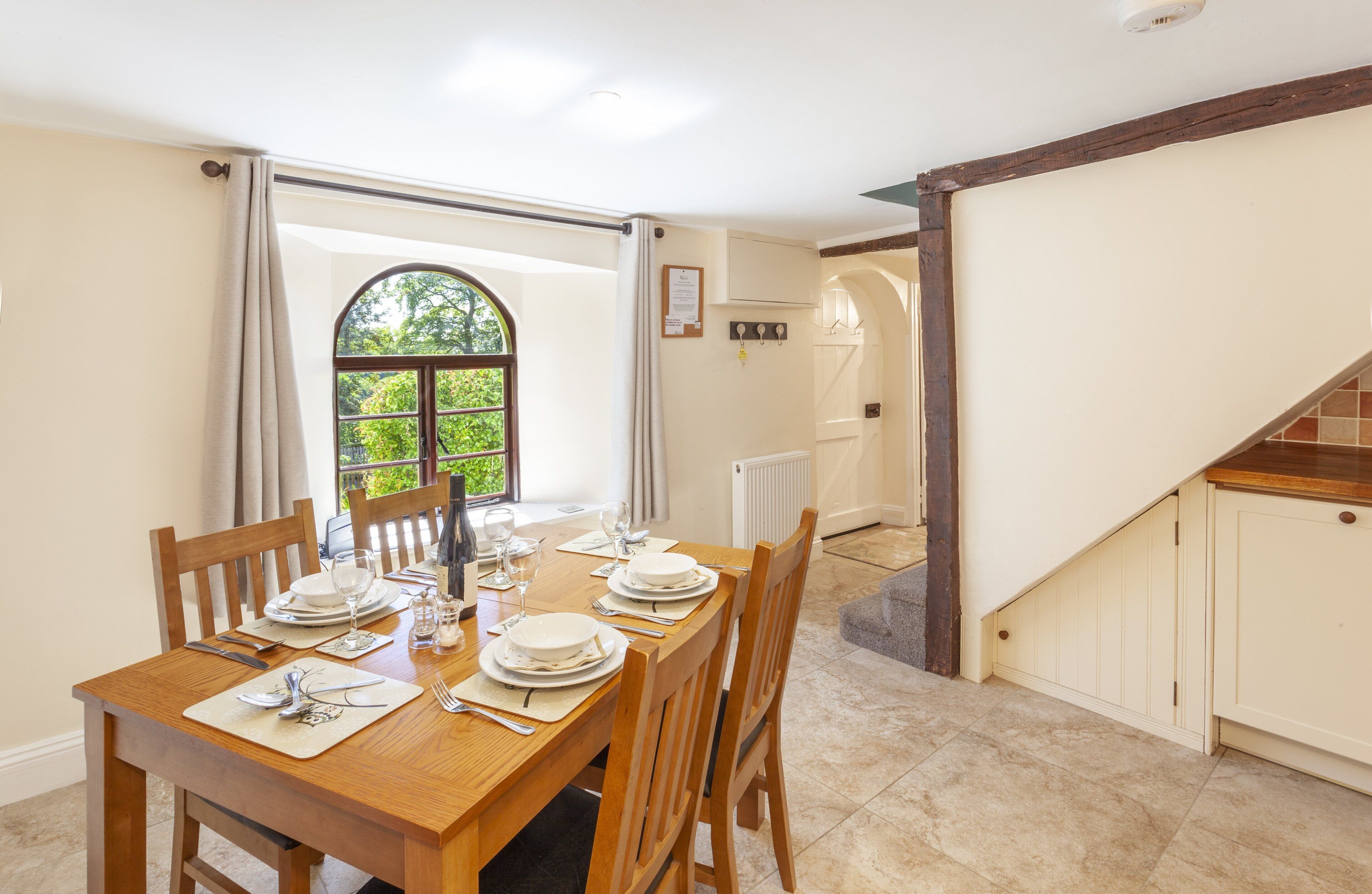A dining area with a wooden table set for four, a window with a view of greenery, and a staircase leading to an upper level.