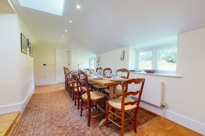 A dining area with a long wooden table set for six, surrounded by wooden chairs with woven seats. A patterned rug covers the floor, and a window provides natural light.