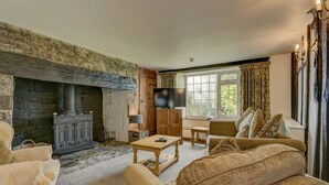 A cozy living room with a stone fireplace, wooden furniture, and a large window with curtains. - The Farmhouse, West Anstey (West Anstey)