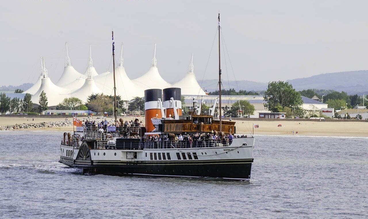 A boat with multiple decks and passengers, moving on water with a beach and white tents in the background.