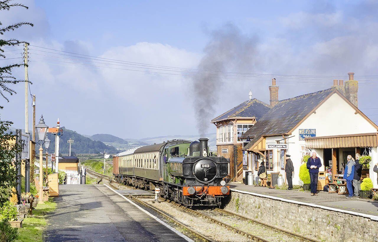 A steam locomotive at a station with a platform, a building labeled 'McGlassine,' and a mountainous backdrop.