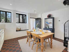 Comedor con mesa de madera dispuesta para cuatro personas, rodeada de sillas. Hay una ventana con vista a la vegetación exterior, un mueble oscuro con estantes y una escalera con barandilla negra.
