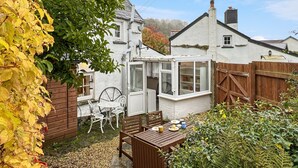 A garden with a wooden table and chairs, a white conservatory, and a white house with a chimney. - Riverside Cottage, Brendon (Nr Lynton)