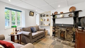 A living room with a fireplace, a sofa, a television, and a window with a view of greenery. - Riverside Cottage, Brendon (Nr Lynton)