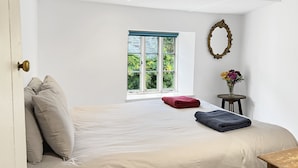 A bedroom with a large bed, a window with a view of greenery, a small table with a vase of flowers, and a mirror on the wall. - Riverside Cottage, Brendon (Nr Lynton)