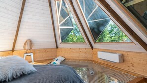 Un dormitorio acogedor en el ático, con techo inclinado, paredes de madera y ventanas amplias que dan a vistas de vegetación.