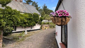 A white building with a stone chimney, a hanging flower basket, and a cobblestone path. - Stables Cottage, Ellicombe (Minehead)