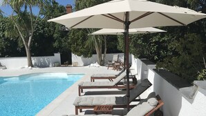 A poolside area with lounge chairs and umbrellas, a clear blue pool, and surrounding greenery. - Stables Cottage, Ellicombe (Minehead)