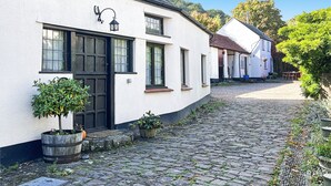 A white building with black-framed windows and a black door, a cobblestone path, and potted plants. - Stables Cottage, Ellicombe (Minehead)