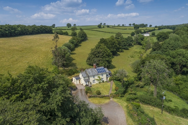 An aerial view of a house with a solar panel roof, surrounded by lush green fields and trees.