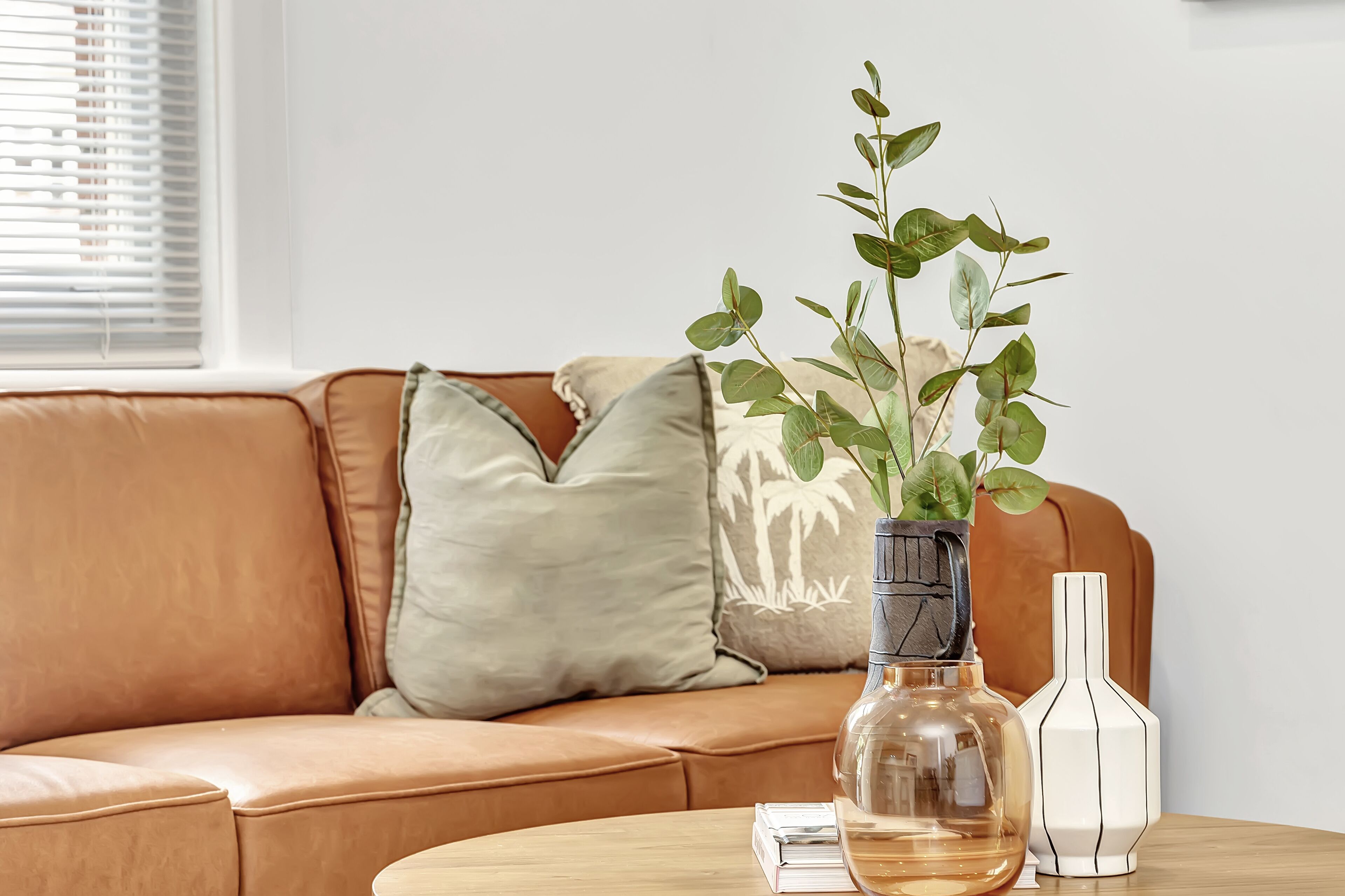 Um sofá de couro cor de bege com uma almofada estampada em branco e verde, um vaso transparente com folhas verdes e um vaso branco com listras pretas sobre uma mesa de madeira.
