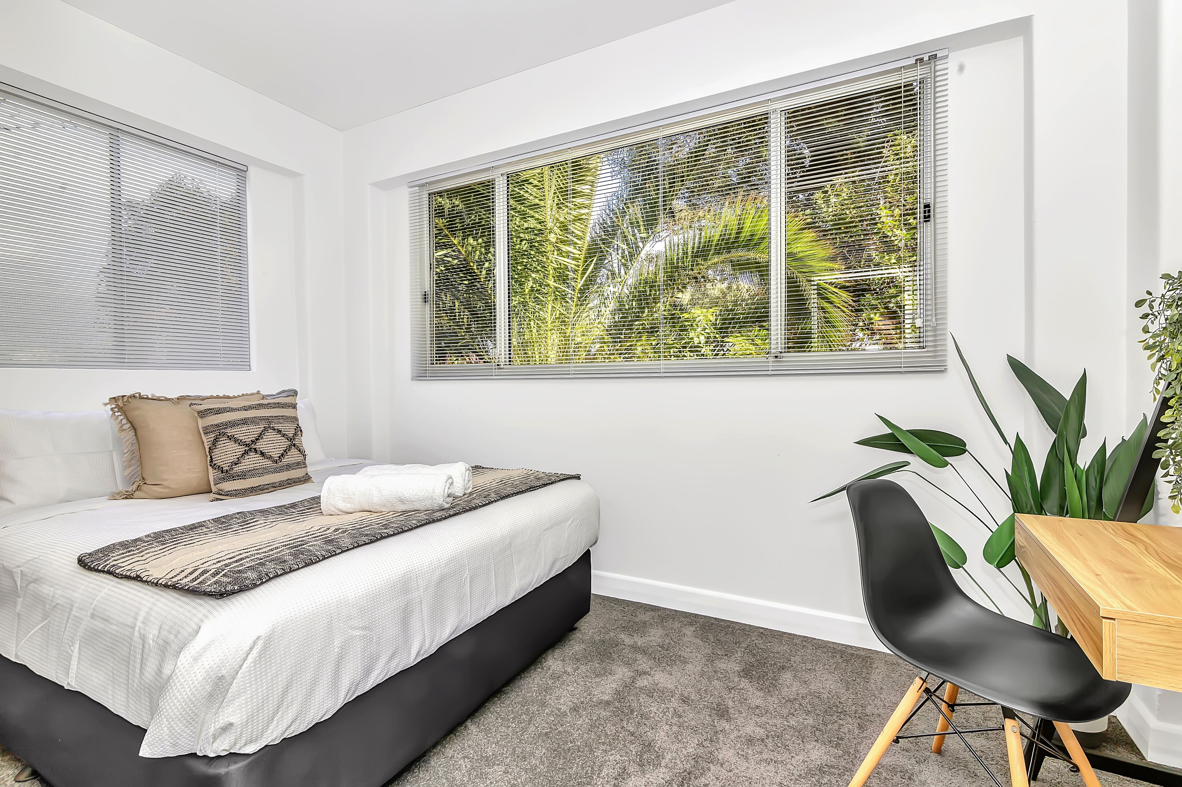 Une chambre à coucher avec un lit, un bureau et une chaise. Il y a une fenêtre avec des stores et une vue sur la verdure à l’extérieur.