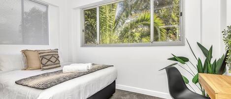 Une chambre à coucher avec un lit, un bureau et une chaise. Il y a une fenêtre avec des stores et une vue sur la verdure à l’extérieur.