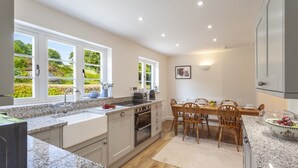 A modern kitchen with granite countertops, a dining area with wooden chairs, and large windows offering a view of greenery. - One Lower Spire Cottage, Liscombe (Nr Dulverton)