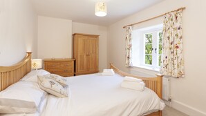 A bedroom with a wooden bed, a wooden wardrobe, a window with floral curtains, and a radiator. - One Lower Spire Cottage, Liscombe (Nr Dulverton)