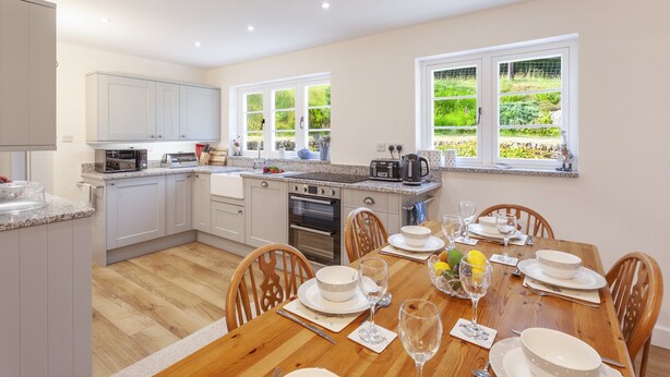 A modern kitchen with white cabinets, a wooden dining table set for six, and a view of a lush garden through large windows. - One Lower Spire Cottage, Liscombe (Nr Dulverton)