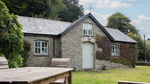 A stone house with a white door and windows, a wooden bench, and a grassy yard.