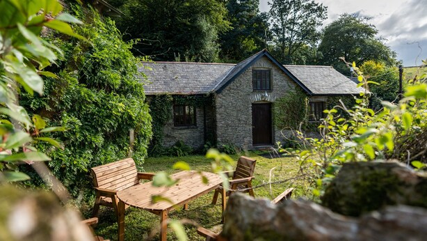 A stone cottage with a thatched roof, surrounded by lush greenery.