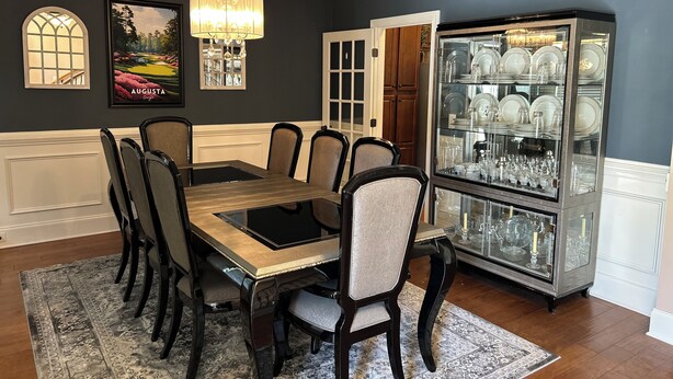 Een eetkamer met een tafel met glazen blad, zes stoelen, een kroonluchter, een vitrinekast met glazen deuren en een ingelijst schilderij aan de muur.