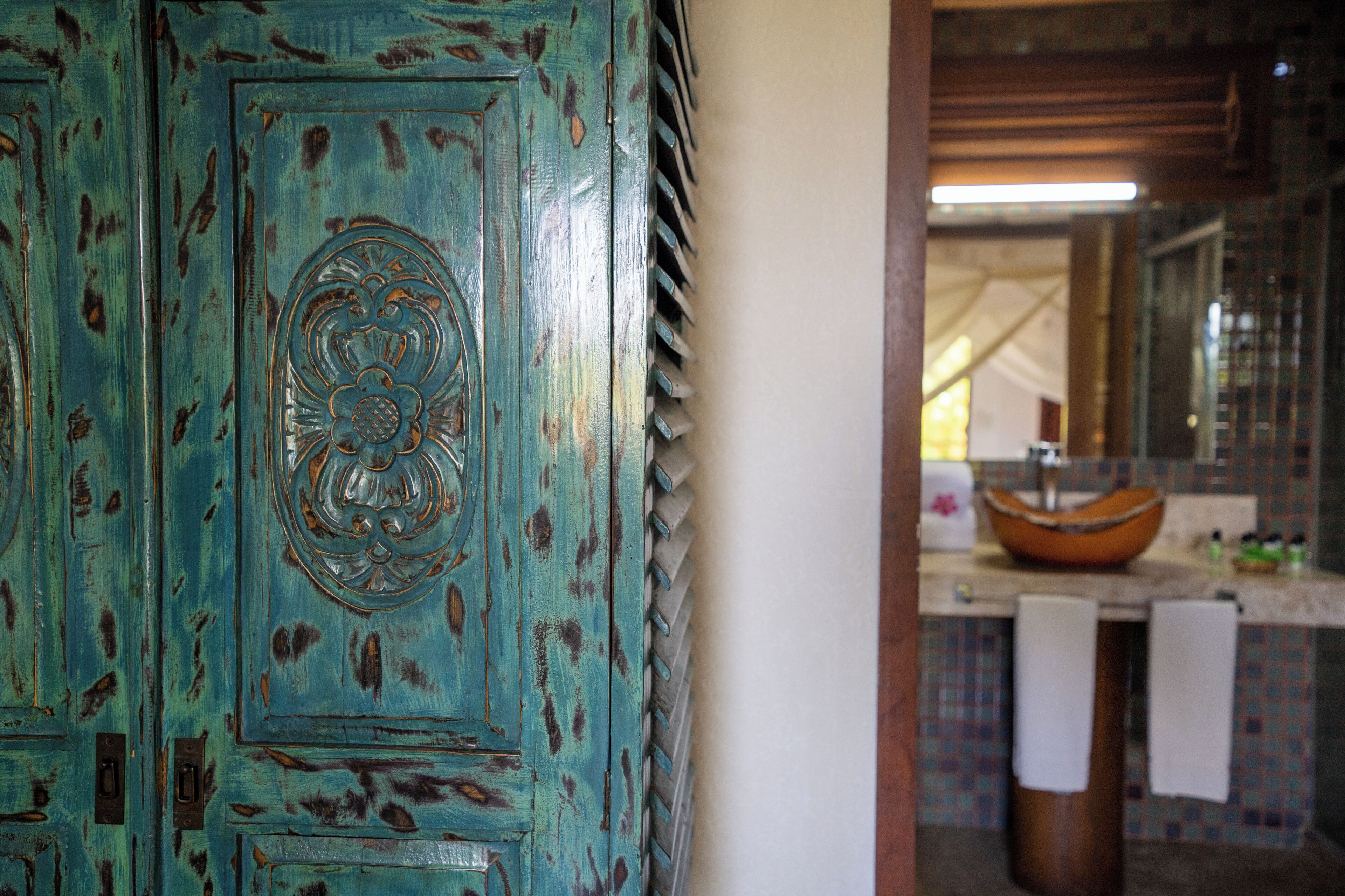 Une porte rustique de couleur turquoise, ornée d’un motif décoratif, qui donne sur une salle de bain dotée d’un grand lavabo rond et d’un mur recouvert de carreaux.
