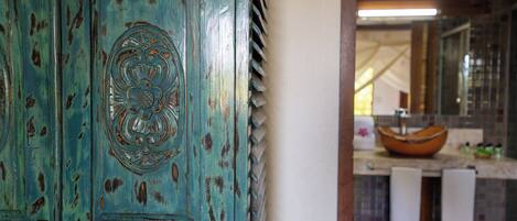 Une porte rustique de couleur turquoise, ornée d’un motif décoratif, qui donne sur une salle de bain dotée d’un grand lavabo rond et d’un mur recouvert de carreaux.