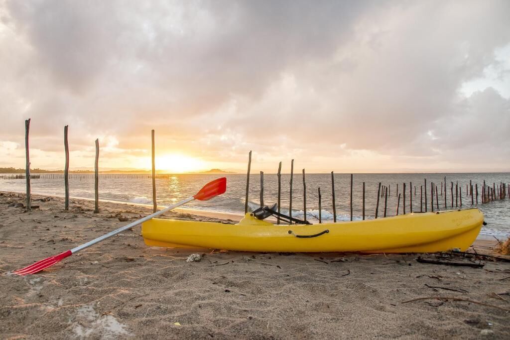 A yellow kayak with a red paddle on a sandy beach with a wooden pier in the background.