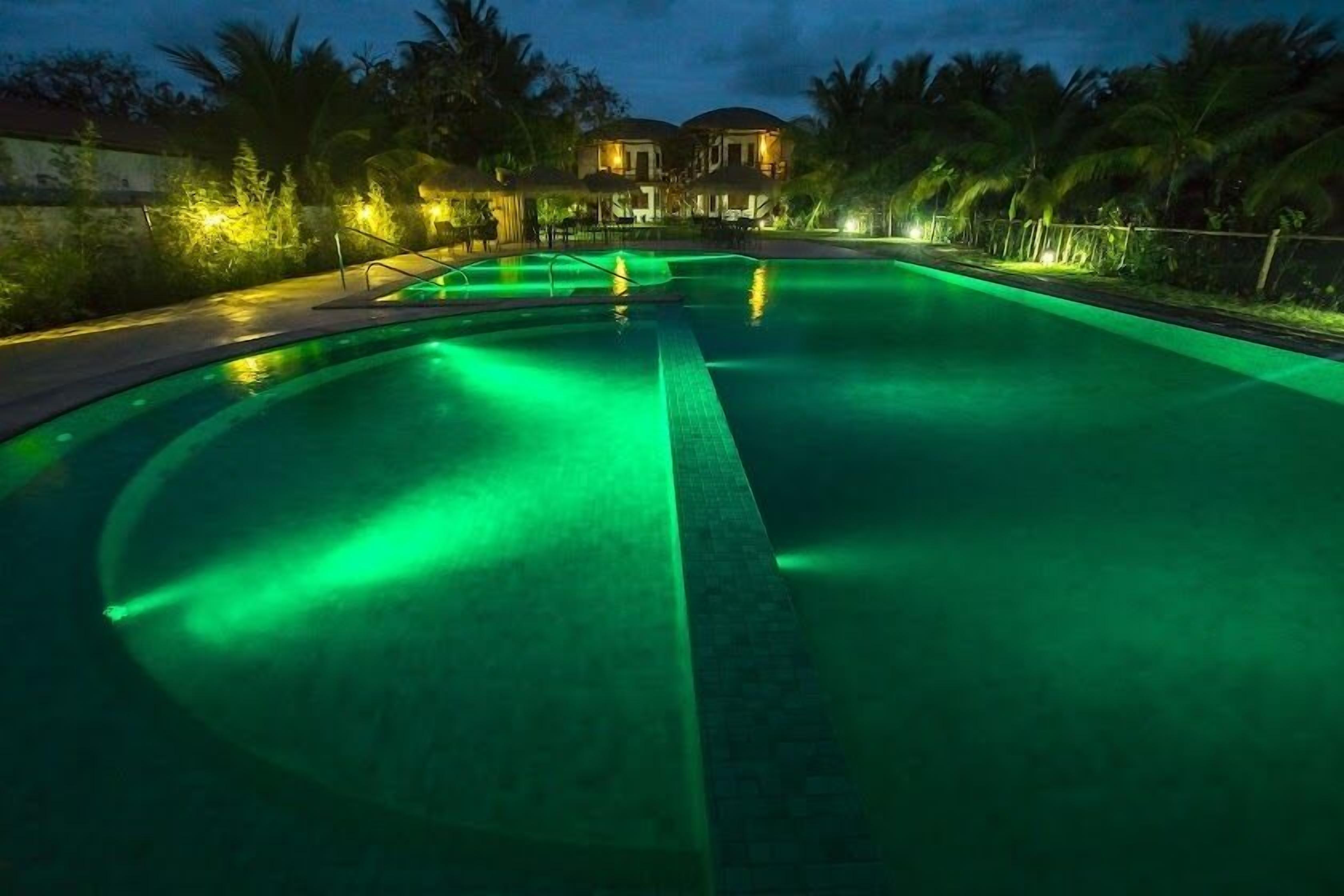 Una piscina iluminada con luz verde, rodeada de palmeras y una casa al fondo.