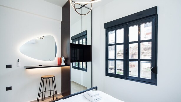 A modern hotel room with a bed, a mirror, a small stool, and a window with a view of buildings outside. - Beautiful Aparment in Psiri-Monastiraki (Athens)