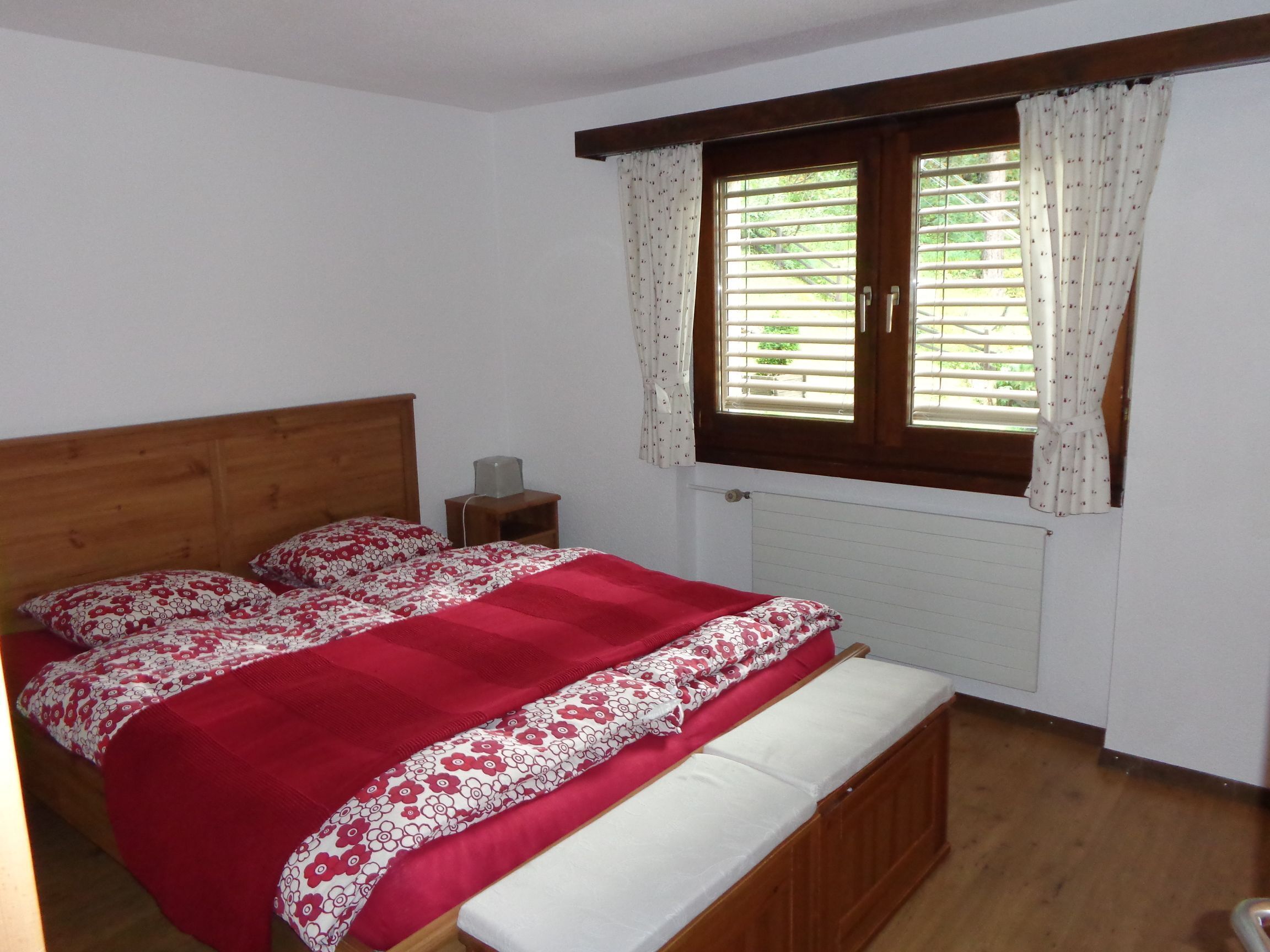 Une chambre à coucher avec un lit en bois, une couvre-lit à motifs rouges et blancs, un banc au pied du lit, une table de chevet avec une lampe et une fenêtre avec des stores et des rideaux.