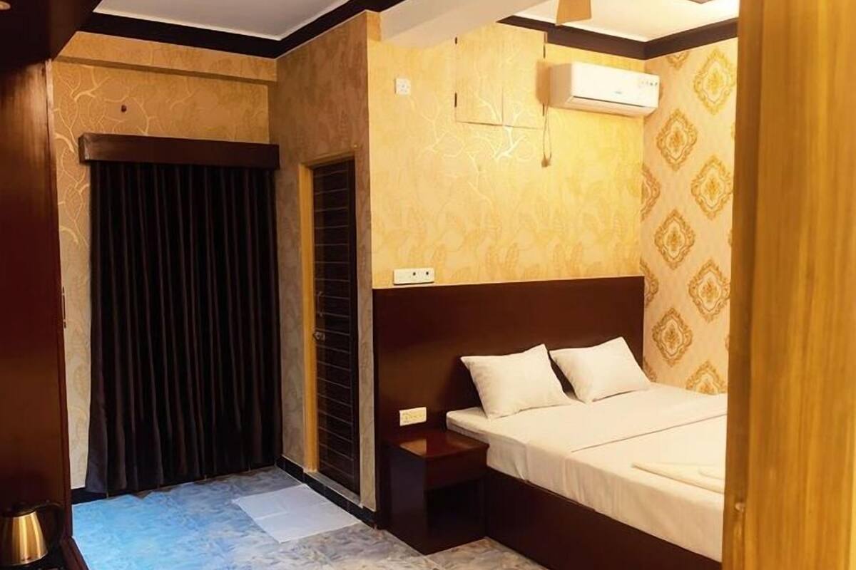 A hotel room with a bed, a nightstand, a window with curtains, and an air conditioning unit.