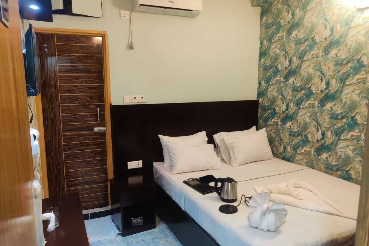 A hotel room with a wooden wardrobe, a bed with white bedding, a kettle, and a remote control on the bed.