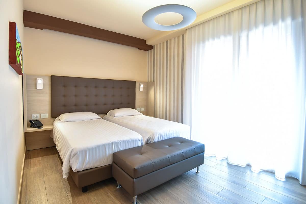 Habitación de hotel con cama doble, mesita de noche pequeña con teléfono, luz en la pared y un ventanal con cortinas.