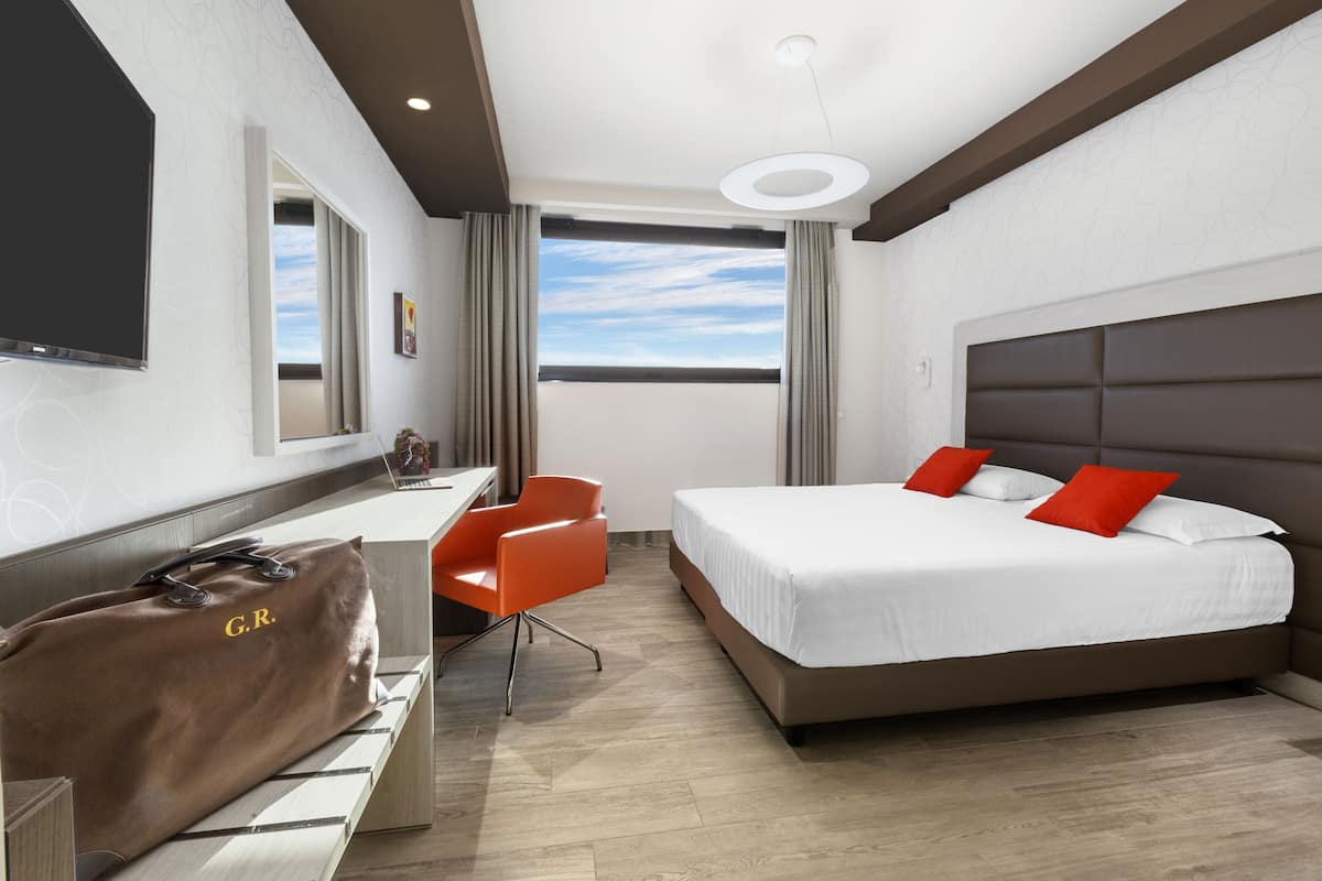 Habitación de hotel con cama, escritorio, silla, ventana grande e iluminación en el techo.