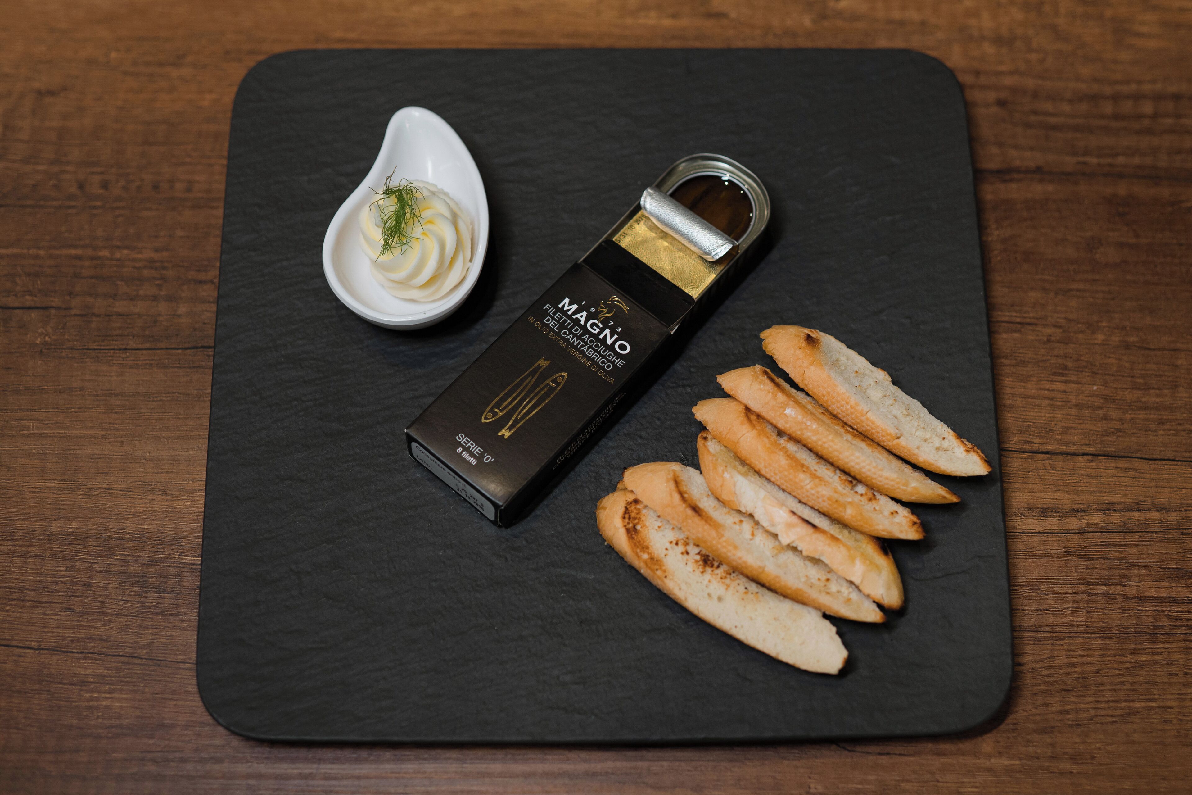 Una pizarra negra con una caja de vino MAGNO Filatura del Carmagnolo, un recipiente blanco pequeño con una sustancia cremosa y varias rebanadas de pan tostado.