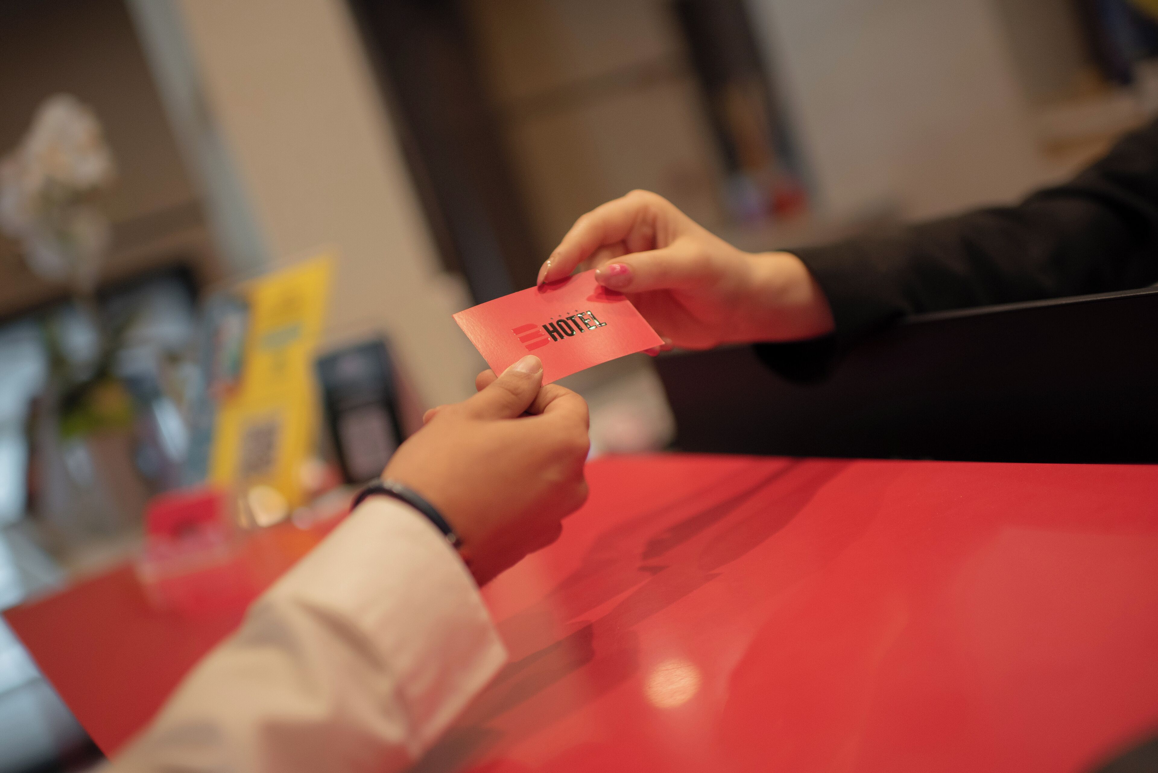 Eine Person übergibt eine rote Karte mit dem Schriftzug 'HOTEL' an eine andere Person am roten Schalter.